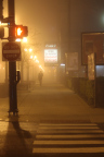 Night time photo of a pedestrian crossing in the foreground, a man looking at his phone in the background, a lit Open 24 Hours
                  Convenient Parking sign above him, and lit streetlights to the left